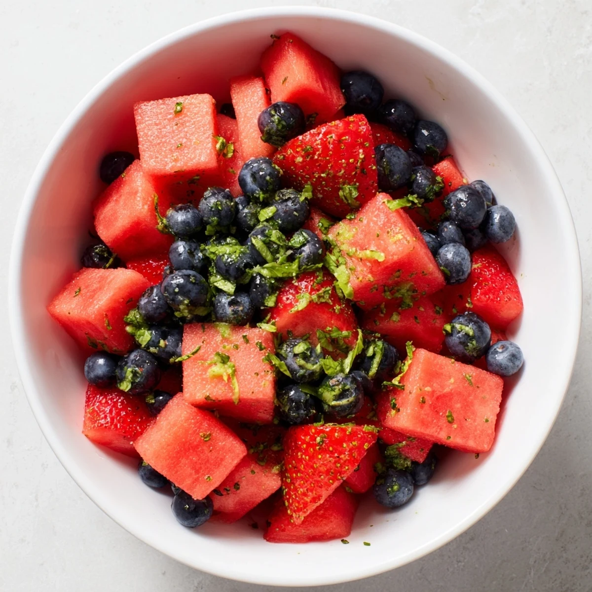 Colorful summer watermelon recipe featuring mixed berries, chopped mint, and citrus-lime honey drizzle on white plate